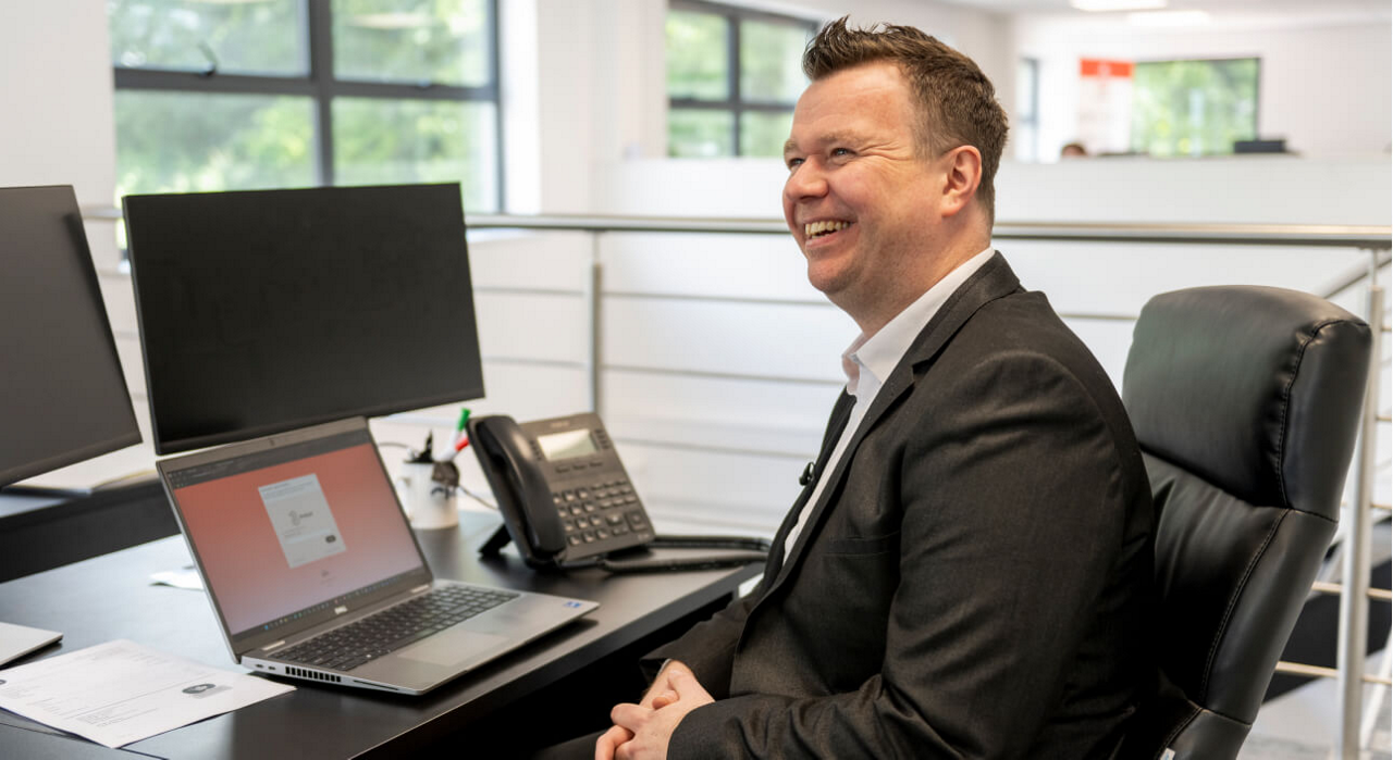 A happy Three Business customer from LV Logistics sits at a modern office desk, smiling while looking at his laptop screen. He is surrounded by dual monitors, a desk phone, and office supplies. The office space has large windows, allowing natural light to fill the area.